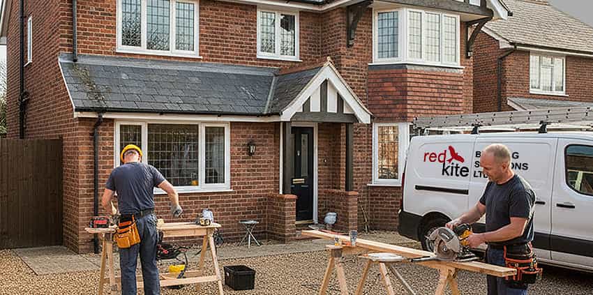 Workmen performing building and maintenance works outside a residential property, using workbenches and tools to carry out repairs.